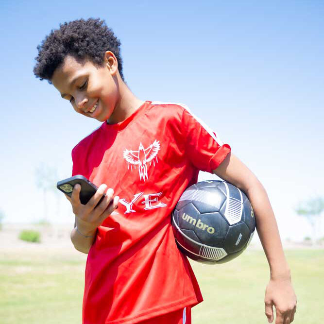 Kid smiling and looking at phone while holding a soccer ball